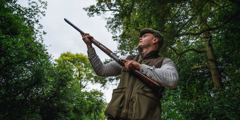Clay shooter standing on a spring shooting ground in bright morning light, wearing layered outdoor clothing and shooting glasses